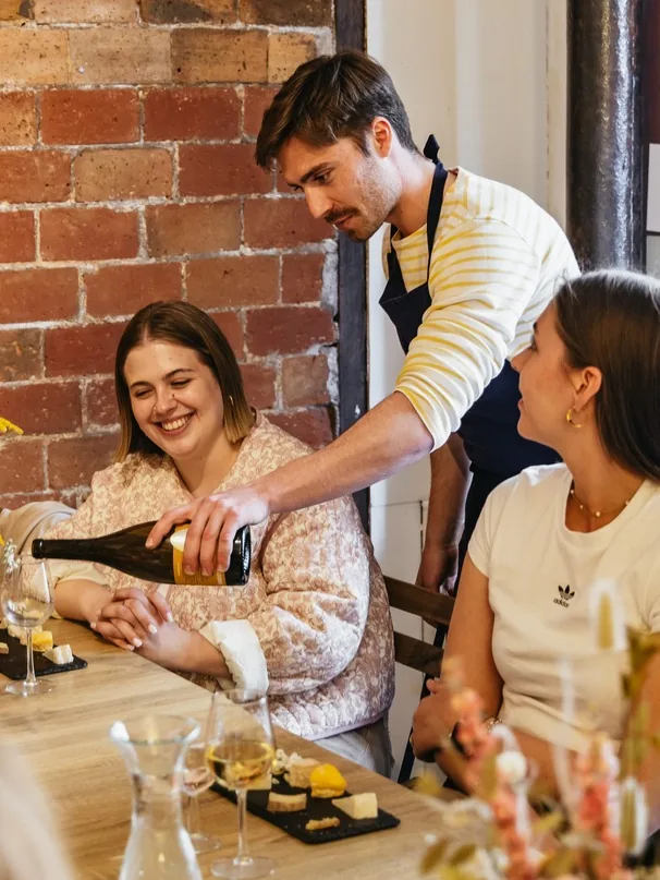Un expert sert un vin blanc à une participante assise, planche de fromages devant elle